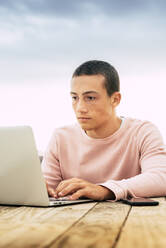 Jugendlicher mit kurzen Haaren, der einen Laptop am Tisch gegen den Himmel benutzt - SIPF02178