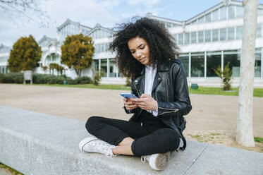 Junge Afro-Frau, die im Schneidersitz auf einer Stützmauer sitzt und ein Smartphone benutzt - KIJF03167