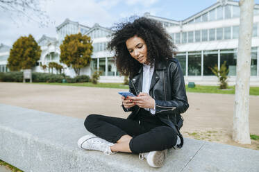 Young afro woman using smart phone while sitting cross-legged on retaining wall - KIJF03167