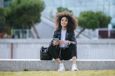 Lächelnde junge Afro-Frau sitzt mit Smartphone und Handtasche auf einer Stützmauer - KIJF03171