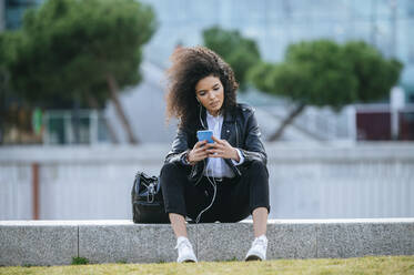 Young woman with afro hairstyle using smart phone while sitting on retaining wall - KIJF03172