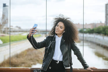 Lächelnde junge Afro-Frau, die ein Selfie macht, während sie sich auf ein Geländer stützt - KIJF03185