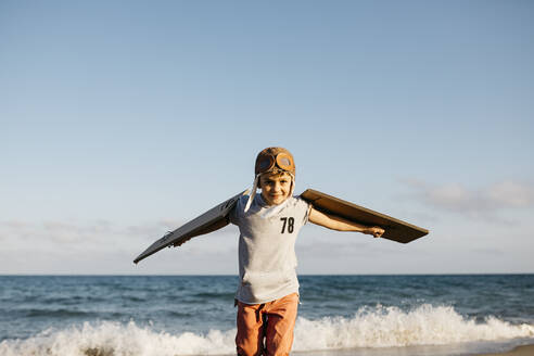 Boy wearing aviator's cap and cardboard wings at beach - JRFF04561