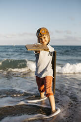 Junge mit Fliegermütze, der am Strand stehend Flügel aus Pappe hält - JRFF04563