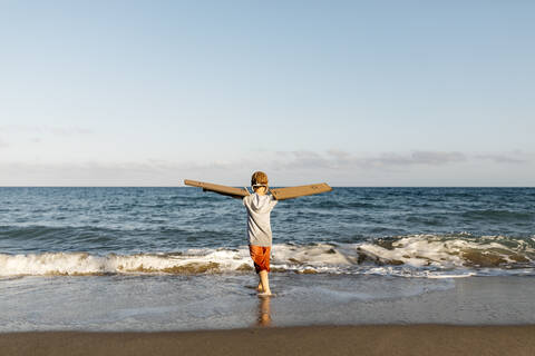 Boy wearing aircraft wings while standing with arms outstretched at beach stock photo