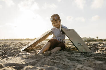 Junge mit Flugzeugflügeln und Mütze auf Sand am Strand sitzend - JRFF04572