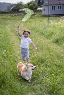Glücklicher Junge mit Schmetterlingsnetz, der mit seinem Hund auf einer Wiese läuft - VPIF02545
