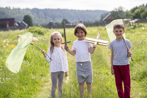 Smiling friends holding butterfly nets and toys while standing on grassy land - VPIF02547