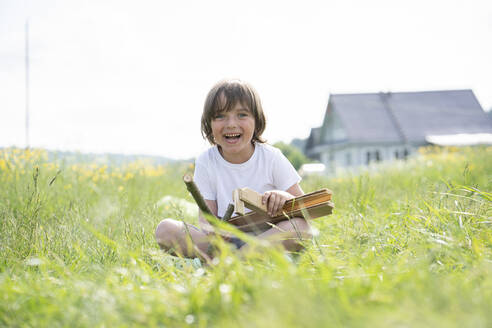 Cheerful cute boy holding model airplane while sitting on grassy land against clear sky - VPIF02550