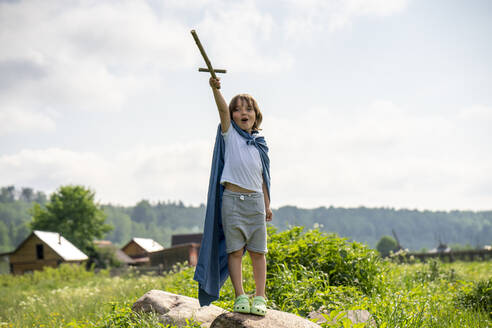 Playful boy wearing cape holding toy sword while standing on rock against sky - VPIF02553