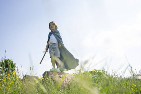 Playful boy wearing cape standing on rock against sky during sunny day - VPIF02554