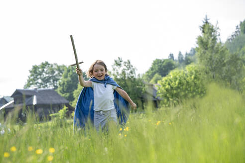 Playful boy wearing cape holding toy sword while running on grassy land against clear sky - VPIF02556