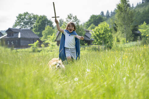 Playful boy wearing cape running with dog on grassy land - VPIF02558