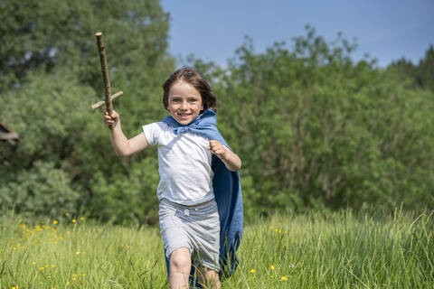 Playful boy wearing cape holding toy sword while running on grassy land stock photo