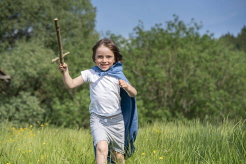 Playful boy wearing cape holding toy sword while running on grassy land - VPIF02559