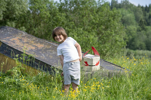 Smiling boy with mask on abandoned boat standing amidst plants in forest - VPIF02570