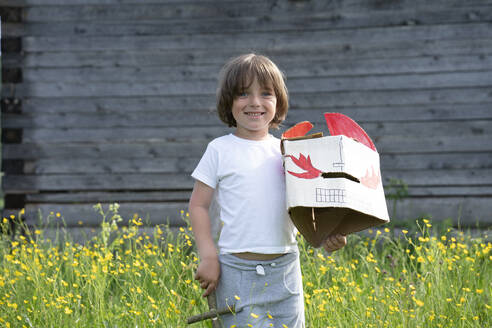 Smiling boy holding mask and toy while standing amidst plants against cottage - VPIF02572