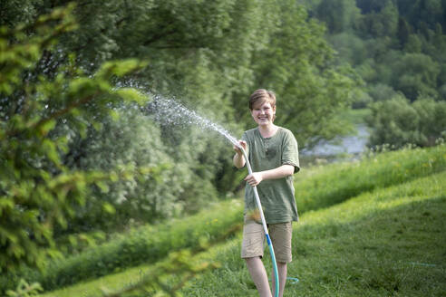 Smiling boy spraying water with hose in forest - VPIF02573