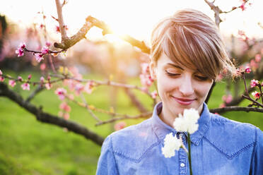 Nahaufnahme einer lächelnden Frau, die bei Sonnenuntergang Blumen im Park betrachtet - SBAF00004