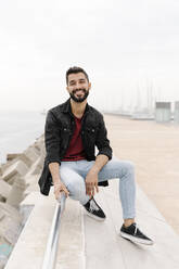 Lächelnder junger Mann auf dem Geländer sitzend gegen den klaren Himmel in der Stadt - RDGF00010