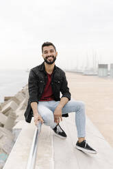 Smiling young man sitting on railing against clear sky in city - RDGF00010