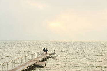 Two people standing on a jetty looking out over water to a pale sunset. - CUF55529