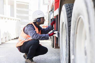 Male engineer wearing hardhat and ear protectors checking undercarriage of a lorry. - CUF55566