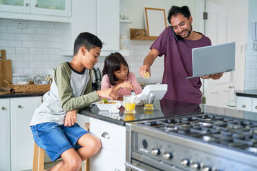 Vater mit Laptop füttert Kinder mit Essen zum Mitnehmen in der Küche - CAIF28269