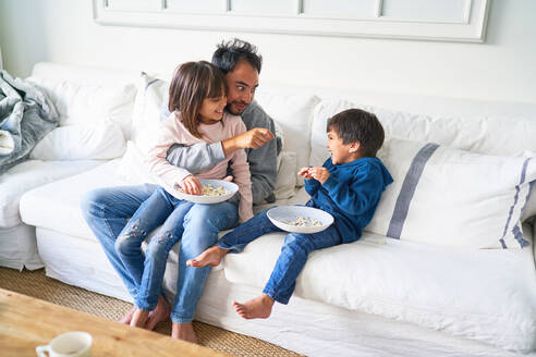 Vater und Kinder essen Popcorn auf dem Wohnzimmersofa - CAIF28274