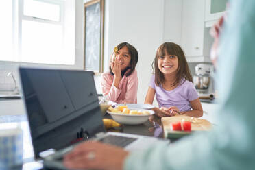Glückliche Schwestern beim Frühstück in der Küche, während die Mutter am Laptop arbeitet - CAIF28329