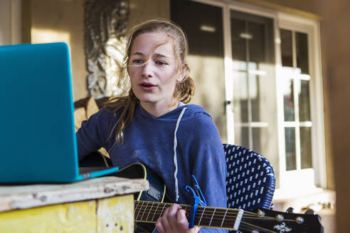 Ein Teenager-Mädchen schaut auf einen Laptop, singt und spielt Gitarre - MINF14606