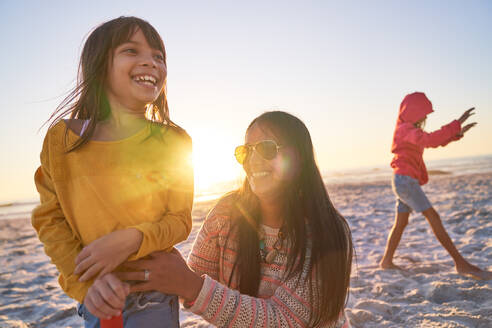 Glückliche Mutter und Tochter spielen am sonnigen Strand bei Sonnenuntergang - CAIF28343