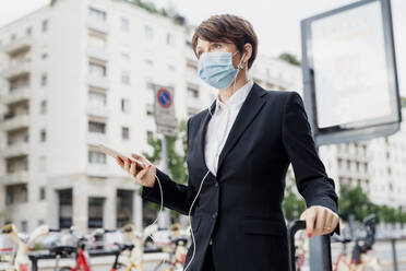 Businesswoman wearing mask holding smart phone while standing in city - MEUF01205