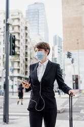 Businesswoman wearing mask holding smart phone while standing with suitcase in city - MEUF01206