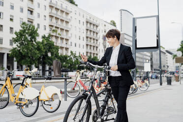 Businesswoman listening music while walking with bicycle on sidewalk in city - MEUF01210