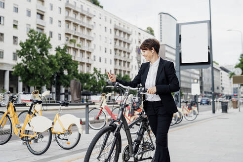 Geschäftsfrau hört Musik, während sie mit dem Fahrrad auf dem Gehweg in der Stadt spazieren geht - MEUF01210