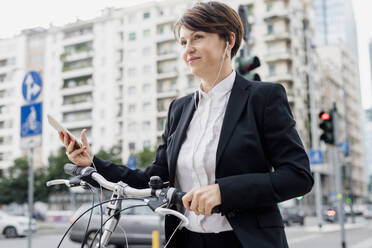 Female professional listening music while standing with bicycle in city - MEUF01213