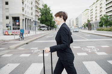 Smiling businesswoman with suitcase crossing road in city - MEUF01221