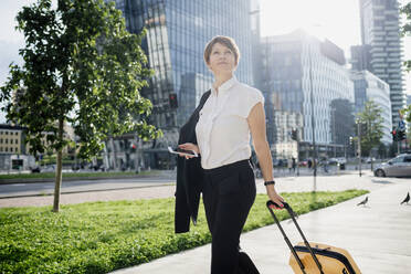 Confident businesswoman holding smart phone and suitcase while walking in city - MEUF01230