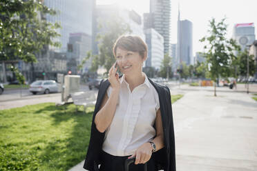 Smiling businesswoman talking over smart phone looking away while standing on sidewalk in city - MEUF01231