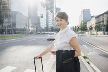 Smiling female professional holding mobile phone and suitcase while standing on road in city - MEUF01236