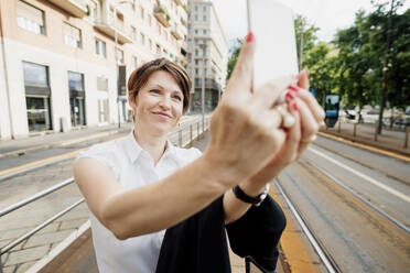 Nahaufnahme einer lächelnden Geschäftsfrau, die ein Selfie mit einem Smartphone in der Stadt macht - MEUF01240