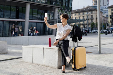 Businesswoman taking selfie with smart phone while sitting by suitcase on seat in city - MEUF01242