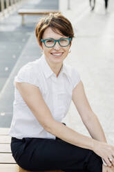 Lächelnde weibliche Fachkraft mit Brille sitzt auf einem Sitz in der Stadt - MEUF01249