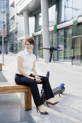Smiling businesswoman with electric push scooter sitting on seat in city - MEUF01252