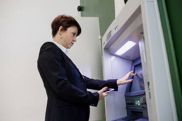 Geschäftsfrau, die ein Smartphone hält und einen Geldautomaten benutzt, während sie an der Wand steht - MEUF01255