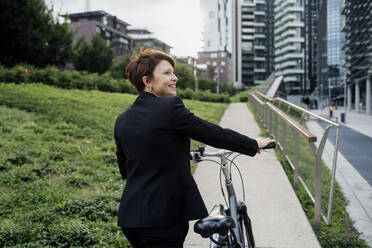 Smiling businesswoman with bicycle walking on footpath in city - MEUF01261