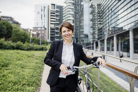 Lächelnde weibliche Fachkraft mit Fahrrad auf dem Fußweg in der Stadt, lizenzfreies Stockfoto