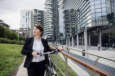 Female professional with bicycle looking away while walking against buildings in city - MEUF01263