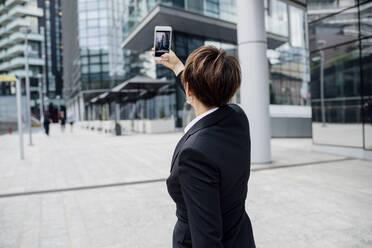 Weiblicher Profi, der ein Selfie mit seinem Smartphone macht, während er vor modernen Gebäuden in der Stadt steht - MEUF01271